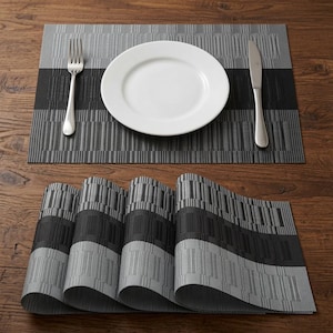 Puede incluir: Un juego de manteles individuales rectangulares con un patrón tejido de rayas horizontales alternas en tonos gris y negro. Un mantel individual está extendido con un plato de cerámica blanca, un tenedor y un cuchillo de metal plateado encima.