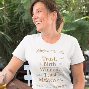 Puede incluir: Una camiseta blanca de manga corta con texto de color dorado que dice "Trust. Birth Women. Trust Midwives.". El texto está enmarcado por delicadas ilustraciones botánicas doradas encima y debajo de las letras, centrado en el pecho.