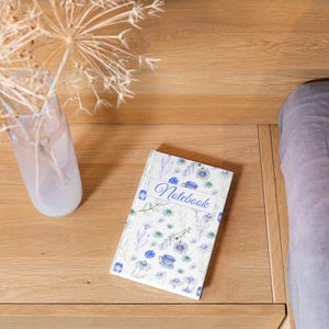 Puede incluir: Un cuaderno rectangular con cubierta blanca que presenta un patrón de acuarela azul con flores, hojas y pequeñas tazas de té. La palabra Notebook está impresa en una tipografía cursiva azul dentro de un marco rectangular central.