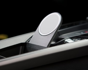 Pode incluir: Um suporte de carregador sem fio magnético circular com acabamento em metal escovado e um disco central cinza claro. O dispositivo está posicionado em ângulo dentro de um console central automotivo escuro, com um design elegante e discreto.