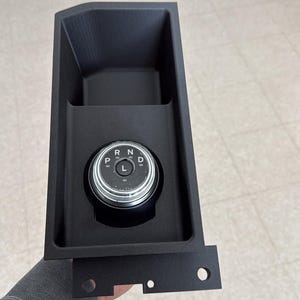 May include: A black automotive gear shift dial assembly featuring a circular knob with a silver metallic rim. The dial displays the letters P, R, N, D, and L in white text. The unit is housed within a rectangular black plastic console frame.