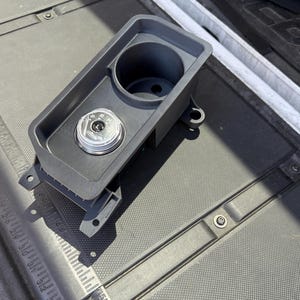 May include: A dark gray automotive center console insert featuring a circular cup holder and a rotary gear shift dial. The dial displays the letters P, R, N, D, and L in white text against a black background with a metallic silver outer ring.