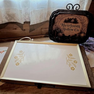 May include: A rectangular white serving tray with a thin gold-colored metal frame. The tray features two symmetrical gold floral sprig designs on the left and right sides. The tray is positioned horizontally on a dark wooden surface.