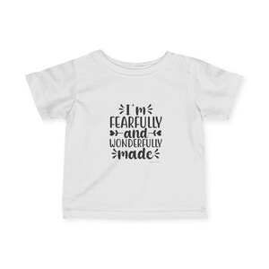Op de afbeelding: Een wit t-shirt met korte mouwen en zwarte tekst in het midden van de borst met de tekst, I'm FEARFULLY and WONDERFULLY made, voorzien van decoratieve krullen en kleine hartvormige iconen geïntegreerd in het typografische ontwerp.