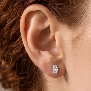 Può includere: Un orecchino a lobo singolo con una pietra preziosa trasparente di forma ovale incastonata in una montatura in metallo color argento lucido. L'orecchino è posizionato sul lobo inferiore dell'orecchio, mettendo in risalto le sfaccettature.