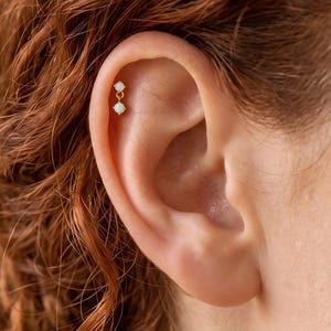 May include: A gold-toned cartilage earring featuring two small, round, iridescent white stones. The top stone is fixed to the post, while the bottom stone hangs from a short, delicate gold chain link, creating a subtle dangling effect on the ear.