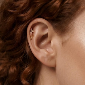 Può includere: Un gioiello per piercing alla cartilagine dell'orecchio color oro, caratterizzato da un intricato design a nodo simmetrico sulla parte superiore, con due delicate catenine pendenti che terminano in piccoli ciondoli lisci a goccia.