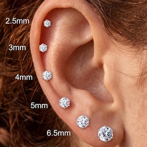 May include: A series of six silver-toned stud earrings with clear round-cut stones, arranged in ascending order of size along the earlobe and cartilage. The sizes are labelled as 2.5mm, 3mm, 4mm, 5mm, 6.5mm, and 7mm, showing the scale of each earring.