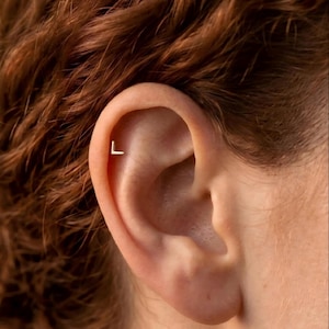 May include: A small, gold-colored metal earring shaped like the capital letter L. The earring is positioned in the upper cartilage area of the ear, featuring a clean, angular design with a polished finish that reflects light against the skin.