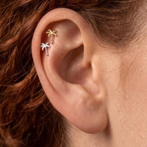 Peut inclure: Deux petites boucles d'oreilles en forme de palmier sont placées sur le cartilage supérieur d'une oreille. L'une est en métal doré et l'autre en métal argenté. Toutes deux présentent des palmes détaillées et un tronc texturé.