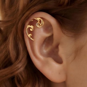 May include: A gold ear cuff featuring two separate, curved, scroll-like metal pieces that wrap around the upper cartilage of the ear. The polished gold finish reflects light, highlighting the smooth, flowing lines of the decorative metal design.