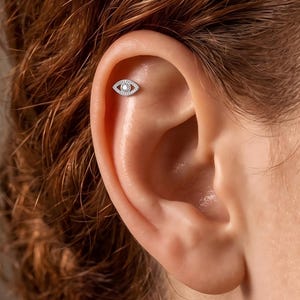 May include: A silver-toned ear stud shaped like an eye, featuring a central clear stone surrounded by a pavé-set border of small clear stones. The jewellery is positioned in the upper cartilage area of the ear, showcasing a detailed eye motif.
