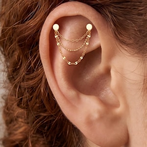 May include: A gold-toned ear cartilage piercing jewellery piece featuring two flat circular studs connected by three delicate, draping chains. The chains vary in texture, with two featuring flat, reflective links and one central chain composed of small beads.