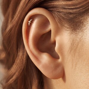 May include: A gold-toned cartilage earring featuring a cluster of four small, clear, marquise and round-cut stones arranged in a vertical, slightly curved line, securely positioned along the upper edge of the ear cartilage.