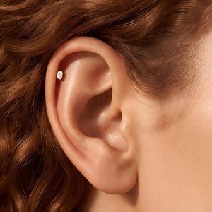 Könnte beinhalten: Ein kleiner, ovaler Ohrstecker mit einem klaren, facettierten Edelstein in einer Metallfassung. Der Ohrstecker befindet sich im oberen Knorpelbereich des Ohrs und weist eine reflektierende Oberfläche auf, die das Licht auf der Haut einfängt.