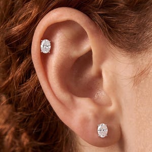 May include: Two silver-toned stud earrings featuring oval-cut clear stones held in four-prong settings. One earring is positioned in the earlobe, while the second earring is placed in the upper cartilage area of the ear, showcasing a matching pair.