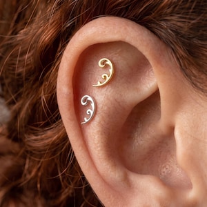 May include: Two curved ear piercings are positioned along the upper cartilage. One piece is finished in a bright gold tone, while the other is finished in a silver tone. Both items feature a delicate, swirling filigree pattern with pointed ends.
