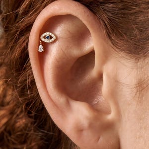 May include: A gold cartilage earring featuring an eye-shaped design encrusted with small clear stones and a central blue stone. A single pear-shaped clear stone hangs from the bottom of the eye motif, creating a delicate and detailed piece of jewelry.