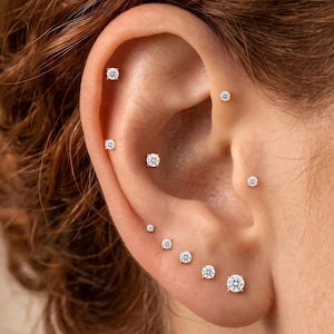 May include: A collection of nine individual stud earrings featuring clear, round-cut stones set in metal prongs. The earrings are arranged in various positions along the ear, including the helix, conch, tragus, and lobe, creating a layered look.