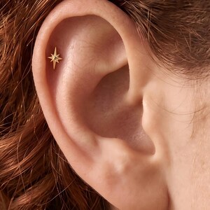 May include: A gold-toned metal ear stud featuring a four-pointed star design with elongated vertical and horizontal rays. The jewelry piece is positioned in the upper cartilage area of the ear, showcasing a polished finish and a sharp, symmetrical shape.