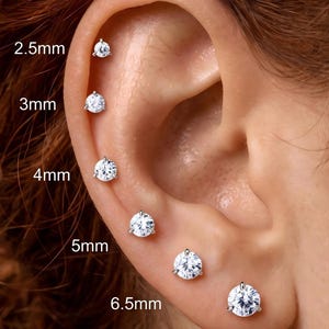 Puede incluir: Una serie de seis pendientes de botón redondos, transparentes y con engaste de garras, dispuestos a lo largo del cartílago y el lóbulo de la oreja. Los tamaños se indican como 2,5 mm, 3 mm, 4 mm, 5 mm, 6,5 mm y 7 mm, mostrando la progresión.