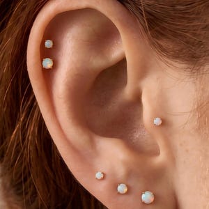 Könnte beinhalten: Ein Set aus sechs runden Opal-Ohrsteckern mit goldfarbenen Fassungen, bestehend aus zwei Steckern im oberen Knorpelbereich, einem im Tragus und drei im Ohrläppchen, die jeweils milchig weiß mit subtilen irisierenden Farbreflexen erscheinen.