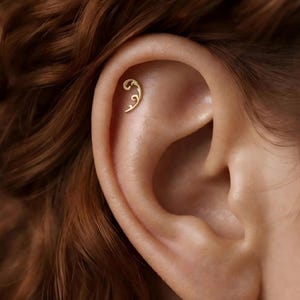 Pode incluir: Um piercing de ouro para a cartilagem da orelha com um design de filigrana curvo e delicado com volutas intrincadas. A joia está posicionada na cartilagem superior, exibindo um acabamento metálico polido e uma forma esguia e elegante.