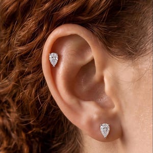 May include: Two silver-toned stud earrings featuring clear, pear-shaped stones. One earring is positioned in the earlobe, while the other is placed in the upper cartilage area. The stones are faceted and held in place by small metal prongs.