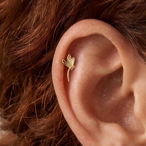 Op de afbeelding: Een goudkleurige oorbel in de vorm van een libel met delicate, langwerpige vleugels en een slank lichaam. Het sieraad is geplaatst op het bovenste kraakbeen van het oor en heeft een gepolijste metalen afwerking met gedetailleerde, symmetrische vleugels.