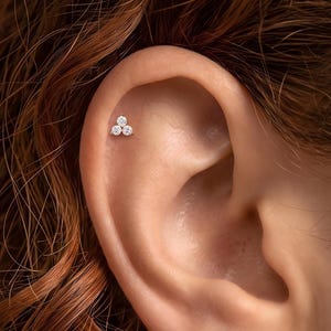 May include: A small, metallic ear stud featuring three round, clear, faceted stones arranged in a triangular cluster. The jewelry is positioned in the upper cartilage area of the ear, showcasing a bright, reflective finish on the setting.