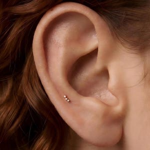 Pode incluir: Um pequeno brinco dourado com três pedras minúsculas, transparentes e de corte redondo, dispostas numa fila vertical. A joia está posicionada na cartilagem da orelha, exibindo um design delicado com um acabamento em metal polido.