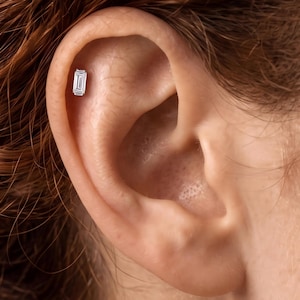 May include: A silver-toned ear piercing stud featuring a rectangular, emerald-cut clear gemstone. The stud is positioned in the upper cartilage area of the ear, showcasing a faceted design that reflects light with a clear, brilliant appearance.