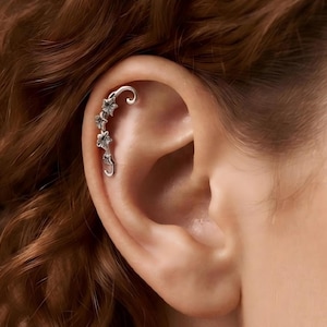 May include: A silver-toned ear cuff featuring a delicate vine design with small, detailed leaves and flowers that follow the natural curve of the upper ear cartilage. The metal has a polished finish with subtle dark accents in the crevices.