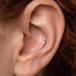 May include: A curved silver-toned ear piercing stud featuring five clear, round-cut stones arranged in a graduated arc. The central stone is the largest, with two smaller stones tapering off on each side, secured in a metal setting within the concha.