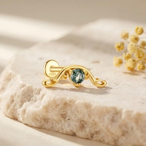 May include: A gold-toned flat back stud earring featuring a curved, ornate metal frame with scrolling details and a single round, moss-green speckled gemstone set in the centre. The post is clearly marked with the text S925 for metal purity.