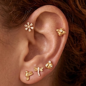 May include: A set of five gold-toned stud earrings featuring nature-inspired designs. The collection includes a flower with baguette-cut stones, a bee with a central stone, a ladybug with two stones, a dragonfly with stone accents, and a bird.