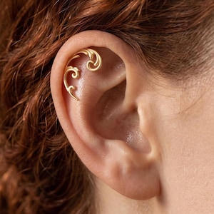 May include: A gold-toned ear cuff featuring a delicate, swirling filigree design that follows the natural curve of the upper ear cartilage. The metal piece has a polished finish and consists of interconnected, elegant scroll-like patterns.