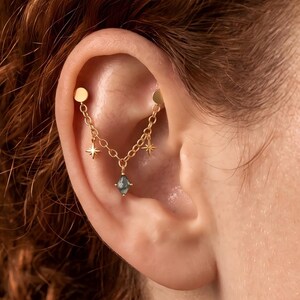 Pode incluir: Uma joia de piercing de orelha em corrente dourada com dois pinos circulares planos conectados por uma corrente delicada. A corrente é adornada com dois pequenos pingentes de estrela de quatro pontas e um pingente central em forma de losango.