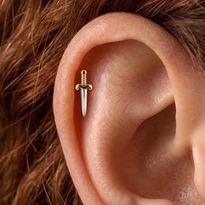 May include: A small earring shaped like a dagger with a gold-colored handle featuring a textured, ridged grip and a smooth, silver-colored blade. The piece is positioned vertically within the upper cartilage area of an ear, showcasing a metallic finish.