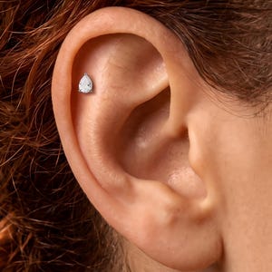 May include: A silver-toned stud earring featuring a clear, pear-shaped gemstone set in a prong setting. The earring is positioned in the upper cartilage area of an ear, showcasing a minimalist design with a single faceted stone that reflects light.