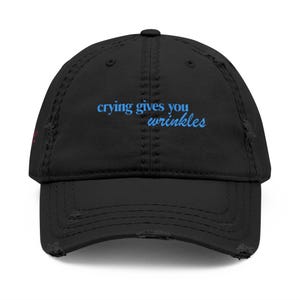 May include: A black distressed baseball cap featuring the embroidered text "crying gives you wrinkles" in light blue on the front panel. The hat has a curved brim with frayed edges and a button on top, showcasing a worn-in aesthetic.