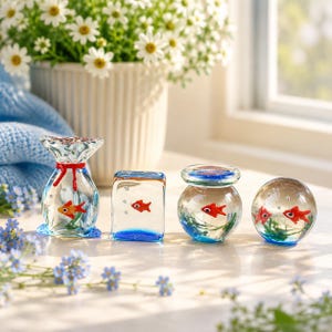 Peut inclure: Quatre ornements en verre transparent contenant des poissons rouges miniatures et des plantes aquatiques vertes. L'ensemble comprend une forme de sac noué, un bloc rectangulaire, un bol rond avec couvercle et une sphère, avec des bulles.