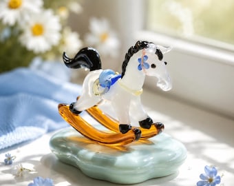 May include: A glass rocking horse figurine featuring a white body, black mane and tail, and amber-coloured rockers. The horse is adorned with a small blue flower detail on its head and a blue saddle, mounted on a smooth, light blue oval base.