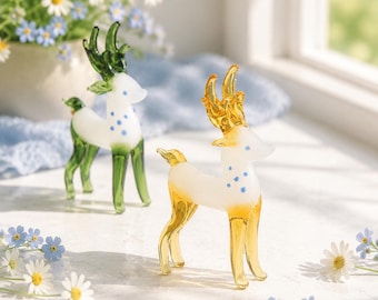 May include: Two glass deer figurines standing on a surface. One deer features a white body with green legs and antlers, while the other has a white body with yellow legs and antlers. Both figurines have small blue spots on their necks.