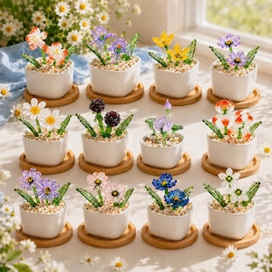 May include: A collection of twelve miniature glass flower arrangements in square white ceramic pots on wooden coasters. Each pot contains small glass flowers in various colors including orange, purple, yellow, white, and blue with green stems.