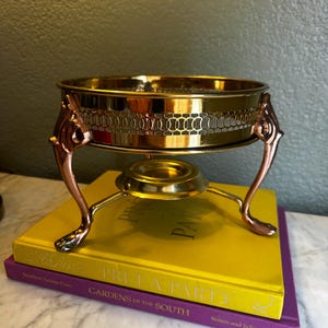 May include: A gold-toned metal chafing dish or warming stand featuring a circular bowl with a decorative lattice-patterned band. The vessel is supported by three curved legs and includes a central circular base for a heat source underneath.