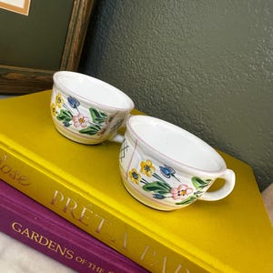 May include: Two white ceramic teacups featuring a hand-painted floral pattern with yellow, pink, and blue flowers, green leaves, and thin pink rim accents. Each cup has a small handle and a rounded body, showcasing a delicate botanical design.
