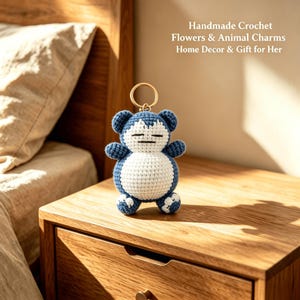 May include: A handmade crochet keychain featuring a blue and white character with a round body, small ears, and closed eyes. The figure is attached to a gold-toned metal ring. Text in the image reads: Handmade Crochet Flowers & Animal Charms Home Decor & Gift for Her.