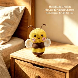 May include: A small handmade crochet bee toy featuring a yellow and brown striped body, white wings, two antennae, black bead eyes, and pink stitched cheeks. The text "Handmade Crochet Flowers & Animal Charms Home Decor & Gift for Her" is visible.