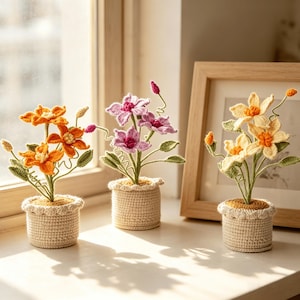 Peut inclure: Trois fleurs crochetées en pot, aux couleurs orange, violette et jaune. Chaque plante présente des tiges et des feuilles vertes, dans un pot en crochet beige avec un bord festonné. Les fleurs ont des pétales détaillés et sont posées.